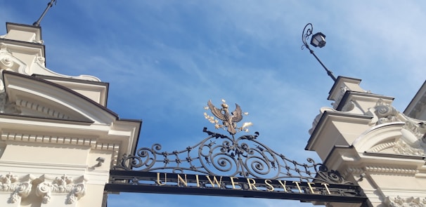 An ornate and wrought iron gate with intricate designs, topped with a golden eagle emblem, stands against a bright blue sky. The structure is flanked by two classical-style columns, suggesting the entrance to a university.