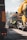 A scene of road construction work with large yellow construction vehicles and workers in high-visibility clothing. Workers are seated on machinery in between the vehicles, which appear to be engaged in paving or maintenance work on a road.