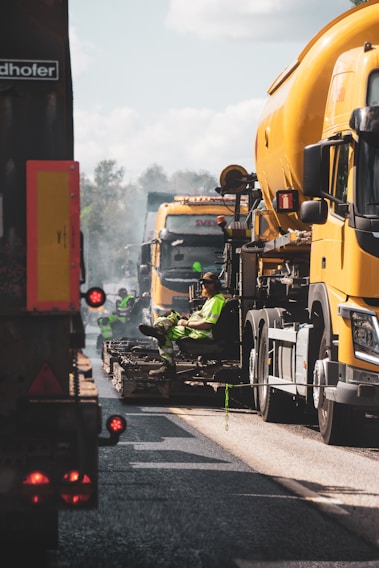 A scene of road construction work with large yellow construction vehicles and workers in high-visibility clothing. Workers are seated on machinery in between the vehicles, which appear to be engaged in paving or maintenance work on a road.