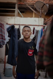 A person stands in an outdoor setting surrounded by hanging clothes. The background consists of a rustic building with exposed beams and a satellite dish. The person has a neutral expression and is wearing a black t-shirt with a red pocket design.