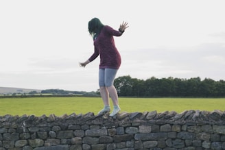 A person with green hair wearing a maroon long-sleeve dress and blue leggings is balancing on a stone wall in a rural landscape. The background features an expansive green field bordered by trees under an overcast sky.