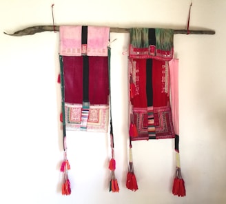 Two colorful hanging textiles are displayed on a wooden rod, featuring intricate patterns and bright tassels at the bottom. The textiles primarily use red, pink, and green hues, with embroidered designs and tassel ornaments.