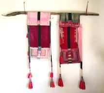 Two colorful hanging textiles are displayed on a wooden rod, featuring intricate patterns and bright tassels at the bottom. The textiles primarily use red, pink, and green hues, with embroidered designs and tassel ornaments.