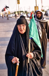An elderly woman in a black abaya, glasses, and headscarf is holding a walking stick and a green flag. She is walking on a busy road with a man in a keffiyeh behind her. There are vehicles and other people in the background, with flags flying from poles.