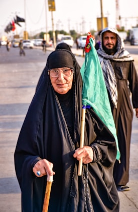 An elderly woman in a black abaya, glasses, and headscarf is holding a walking stick and a green flag. She is walking on a busy road with a man in a keffiyeh behind her. There are vehicles and other people in the background, with flags flying from poles.