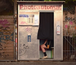 A photo booth with a partially visible person sitting inside, with only their legs and feet visible under a black curtain. The exterior of the booth is metallic with graffiti and stickers. A cartoon drawing of a snail is visible on the booth's surface. The background contains a wire fence and more graffiti.