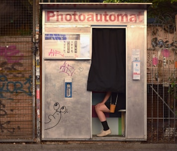 A photo booth with a partially visible person sitting inside, with only their legs and feet visible under a black curtain. The exterior of the booth is metallic with graffiti and stickers. A cartoon drawing of a snail is visible on the booth's surface. The background contains a wire fence and more graffiti.