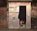 A photo booth with a partially visible person sitting inside, with only their legs and feet visible under a black curtain. The exterior of the booth is metallic with graffiti and stickers. A cartoon drawing of a snail is visible on the booth's surface. The background contains a wire fence and more graffiti.