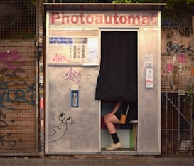 A photo booth with a partially visible person sitting inside, with only their legs and feet visible under a black curtain. The exterior of the booth is metallic with graffiti and stickers. A cartoon drawing of a snail is visible on the booth's surface. The background contains a wire fence and more graffiti.