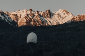 A person wearing a white hooded jacket is standing in front of a rugged mountain range covered with snow. The peaks are sharply defined against a clear sky, and the foreground is a dark silhouette of the person observing the scene.
