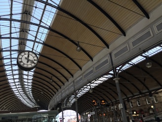 A spacious train station platform featuring an arched glass and metal ceiling. The clock is prominently suspended, displaying the time. Multiple beams support the structure, with hanging lamps and overhead signs visible. The platform area appears tidy and well-maintained.