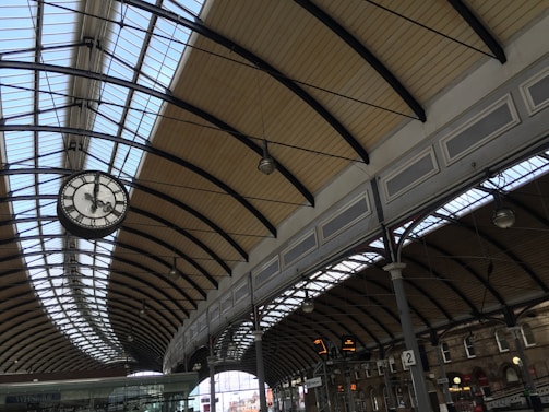 A spacious train station platform featuring an arched glass and metal ceiling. The clock is prominently suspended, displaying the time. Multiple beams support the structure, with hanging lamps and overhead signs visible. The platform area appears tidy and well-maintained.