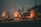 Three individuals are performing a ritual with brass lamps, emitting multiple flames, amidst swirling smoke. They are dressed in traditional attire, adding a sense of ceremony against a twilight backdrop.