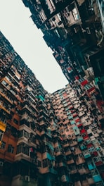 A dense and towering group of residential buildings with a variety of brightly colored facades. The buildings appear to form a narrow alleyway-like space with their sheer height creating an imposing and somewhat chaotic visual. There are numerous windows, air conditioning units, and laundry hanging from lines, indicating an urban lifestyle.