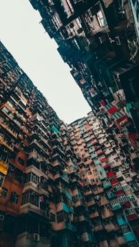 A dense and towering group of residential buildings with a variety of brightly colored facades. The buildings appear to form a narrow alleyway-like space with their sheer height creating an imposing and somewhat chaotic visual. There are numerous windows, air conditioning units, and laundry hanging from lines, indicating an urban lifestyle.