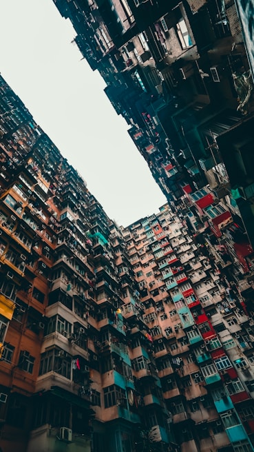 A dense and towering group of residential buildings with a variety of brightly colored facades. The buildings appear to form a narrow alleyway-like space with their sheer height creating an imposing and somewhat chaotic visual. There are numerous windows, air conditioning units, and laundry hanging from lines, indicating an urban lifestyle.