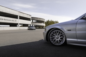 A silver car with sporty rims is prominently featured in the foreground. In the background, there's a parking structure with a concrete design, displaying the text 'LKW-Wartezone max. 2h'. Another car is positioned further back, near the structure. Trees are visible in the distance under a clear sky.