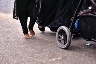 A child is walking barefoot on a paved surface next to a stroller. The child is wearing black clothing, and the stroller has large, visible wheels with some dirt on them. Part of the stroller is also black, with some colorful fabric visible inside.