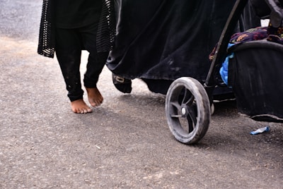 A child is walking barefoot on a paved surface next to a stroller. The child is wearing black clothing, and the stroller has large, visible wheels with some dirt on them. Part of the stroller is also black, with some colorful fabric visible inside.