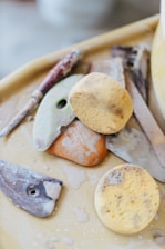 A collection of pottery tools, including sponges, ribs, and carving tools, are arranged on a table. The tools appear well-used, with clay residue visible on the surfaces. The image has a soft focus and a neutral background.