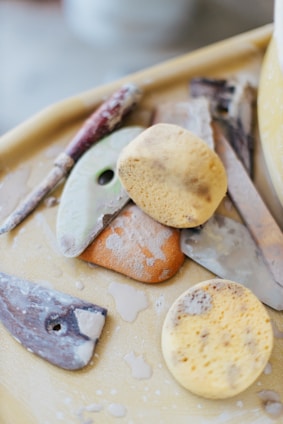 A collection of pottery tools, including sponges, ribs, and carving tools, are arranged on a table. The tools appear well-used, with clay residue visible on the surfaces. The image has a soft focus and a neutral background.