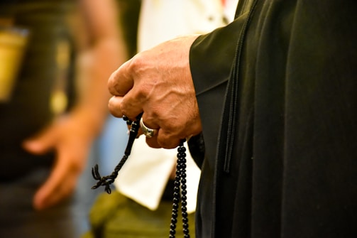 A close-up of a person's hand holding a rosary or prayer beads, highlighting the texture and details of the beads and the hand. The background is blurred, drawing focus to the hand and the beads.