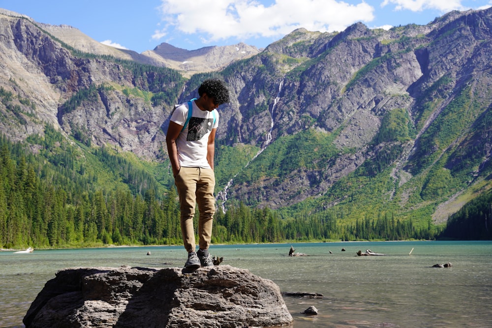 man in white t-shirt and brown pants standing on rock near body of water during