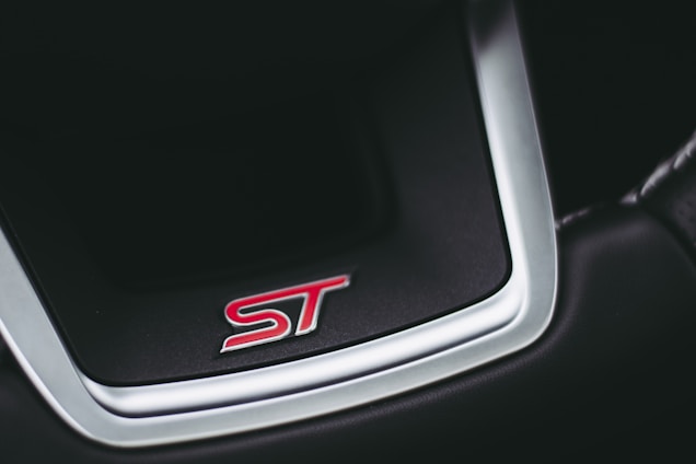 A close-up of a car steering wheel emblem featuring the letters 'ST' in red against a black background, bordered by a shiny metallic frame.