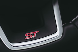 A close-up of a car steering wheel emblem featuring the letters 'ST' in red against a black background, bordered by a shiny metallic frame.