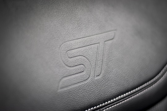The image features a close-up view of a leather texture with embossed letters 'ST'. The surface is detailed and finely textured, highlighting the stitching along the edge.