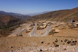 A rural village is nestled in a mountainous terrain with winding roads connecting small, flat-roofed buildings. Sparse vegetation covers the hillsides, and a herd of goats is grazing in the foreground.