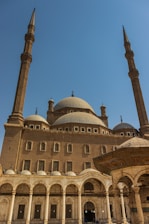 A large, ornate mosque with prominent domes and tall minarets stands under a clear blue sky. The structure features intricate architectural details, including arched entrances and decorative patterns. The mosque exudes an air of grandeur and historical significance.