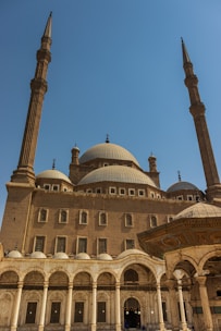 A large, ornate mosque with prominent domes and tall minarets stands under a clear blue sky. The structure features intricate architectural details, including arched entrances and decorative patterns. The mosque exudes an air of grandeur and historical significance.