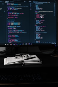 A dimly lit workspace featuring a large monitor displaying lines of colorful code, suggesting a programming environment. In front of the monitor, a notebook with handwritten notes, a pair of glasses, and a pen are neatly placed, indicating an engaged working session.