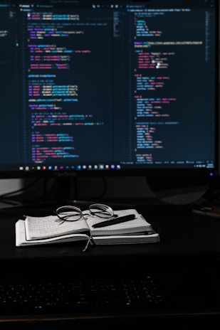 A dimly lit workspace featuring a large monitor displaying lines of colorful code, suggesting a programming environment. In front of the monitor, a notebook with handwritten notes, a pair of glasses, and a pen are neatly placed, indicating an engaged working session.