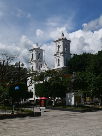 A plaza with a prominent white church featuring two bell towers under a partly cloudy sky. Surrounding the church, there are green trees and a statue on a pedestal in the foreground. A poster with an image of a colorful skull under a pink umbrella is also visible.