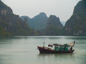 A traditional, weathered fishing boat floats gently on calm waters, surrounded by steep, lush green limestone islands. The sky is overcast, casting a muted light over the tranquil scene. The boat features a small cabin and visible laundry hanging to dry, contributing to a sense of daily life and simplicity.