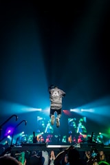 A person wearing a jacket with 'ANOMALY' jumps energetically on a stage during a concert. Bright colorful lights and screens displaying graphics create a vibrant atmosphere. The audience is visible at the bottom, capturing the event with their phones.
