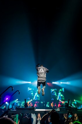A person wearing a jacket with 'ANOMALY' jumps energetically on a stage during a concert. Bright colorful lights and screens displaying graphics create a vibrant atmosphere. The audience is visible at the bottom, capturing the event with their phones.