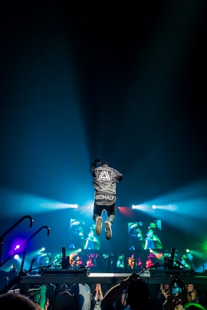 A person wearing a jacket with 'ANOMALY' jumps energetically on a stage during a concert. Bright colorful lights and screens displaying graphics create a vibrant atmosphere. The audience is visible at the bottom, capturing the event with their phones.