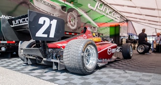 A red race car with the number 21 is parked on a checkered floor inside a tented area. Juncos Racing branding is visible on both the car and the surrounding area. Two people are seated in the background near stacks of tires. A large vehicle wrap with racing imagery is displayed prominently behind the car.