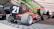 A red race car with the number 21 is parked on a checkered floor inside a tented area. Juncos Racing branding is visible on both the car and the surrounding area. Two people are seated in the background near stacks of tires. A large vehicle wrap with racing imagery is displayed prominently behind the car.
