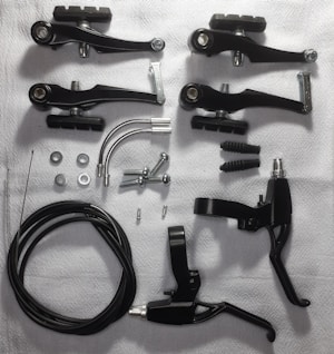 Various bicycle brake components are arranged on a white surface. These include black brake levers, brake arms, cables, screws, washers, and metallic arcs.