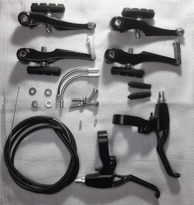 Various bicycle brake components are arranged on a white surface. These include black brake levers, brake arms, cables, screws, washers, and metallic arcs.