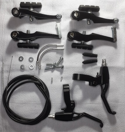 Various bicycle brake components are arranged on a white surface. These include black brake levers, brake arms, cables, screws, washers, and metallic arcs.