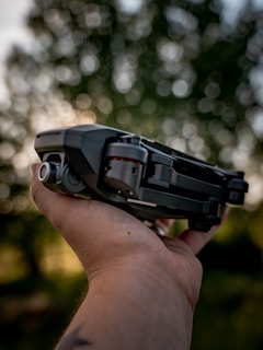 A person's hand holds a compact, folding drone against a backdrop of blurred, lush green foliage. The drone features a sleek black design with visible propellers and camera lens. The background is softly out of focus, creating a bokeh effect.