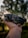 A person's hand holds a compact, folding drone against a backdrop of blurred, lush green foliage. The drone features a sleek black design with visible propellers and camera lens. The background is softly out of focus, creating a bokeh effect.