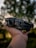 A person's hand holds a compact, folding drone against a backdrop of blurred, lush green foliage. The drone features a sleek black design with visible propellers and camera lens. The background is softly out of focus, creating a bokeh effect.