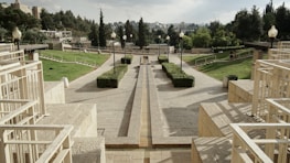 A well-maintained landscaped park featuring a wide stone pathway lined with lamp posts and hedges. The area is surrounded by green lawns and various trees, with beige stone structures and railings on both sides. The skyline consists of trees and distant city structures under a partly cloudy sky.