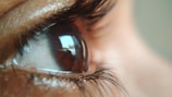 A close-up view of a human eye with detailed focus on the eyelashes and the reflection in the pupil. The reflections show a blurred, abstract pattern, suggesting a distorted view of an environment.